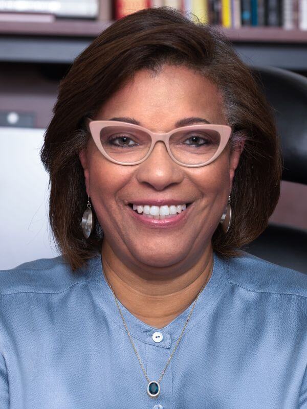 Loretta Parham wearing a blue top, light color glassses, earrings, and a necklace, looking directly into the camera.